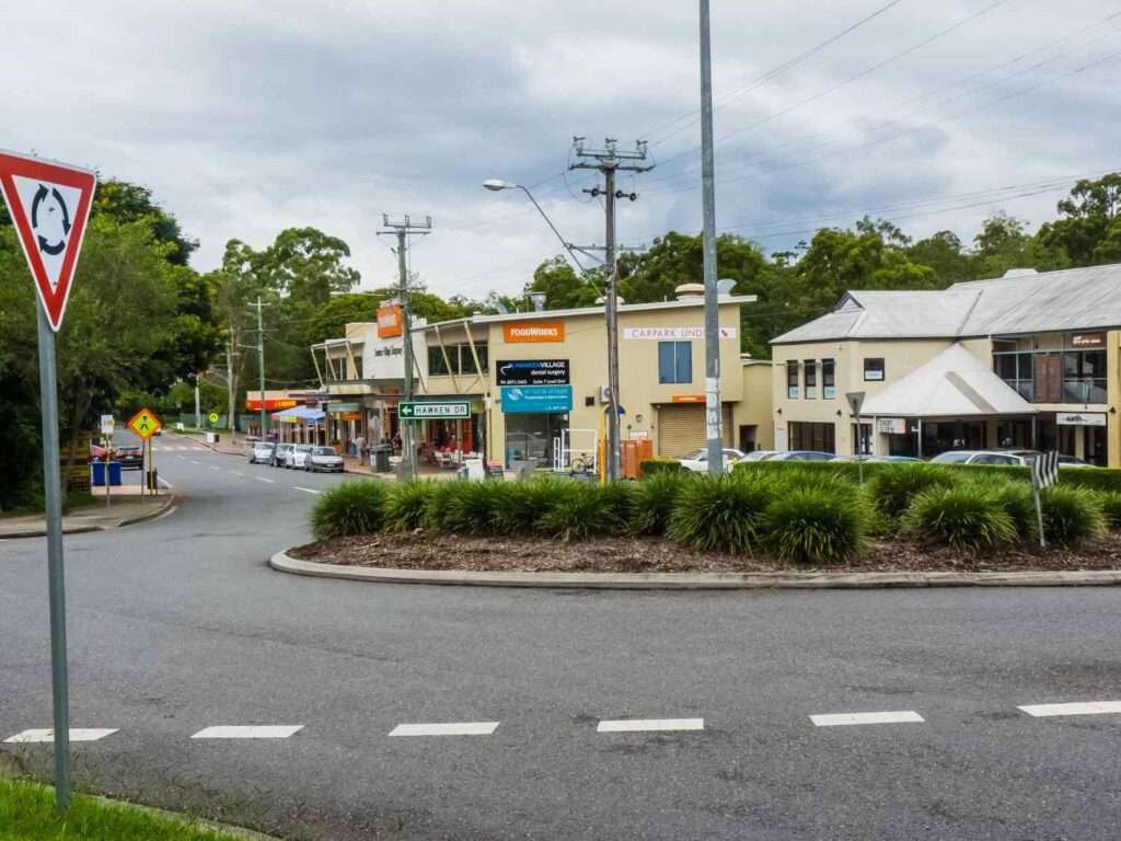 Shops and local businesses along Hawken Drive in St Lucia, Brisbane, with signage, parked cars, and surrounding greenery under an overcast sky.