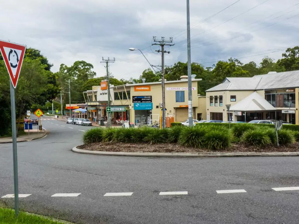 Shops and local businesses along Hawken Drive in St Lucia, Brisbane, with signage, parked cars, and surrounding greenery under an overcast sky.