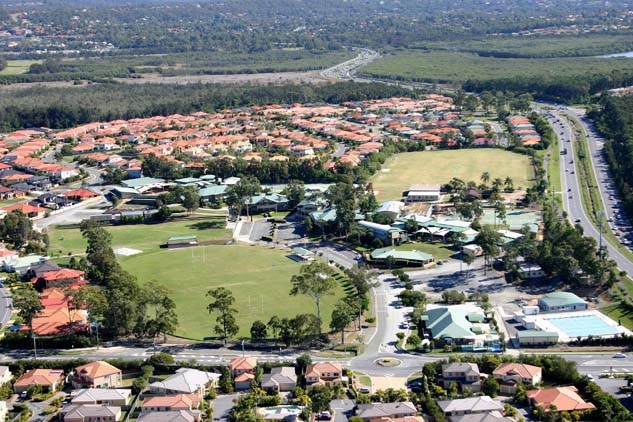 Alt text: Aerial view of suburban homes, schools, and open playing fields in Arundel, Gold Coast, with surrounding bushland and highway access visible in the background.