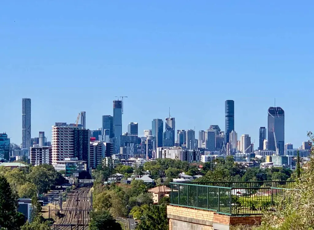 Skyline view of Brisbane CBD seen from Taringa, featuring city skyscrapers, train tracks, and leafy suburban rooftops in the foreground.