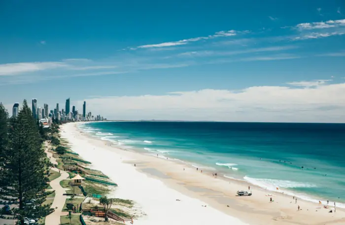 A scenic coastal view from Miami Beach, Gold Coast, looking north towards Surfers Paradise, showcasing white sand, turquoise water, and the distant skyline of high-rise buildings under a bright blue sky.