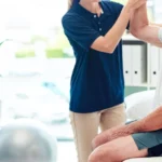 Warm, professional image of a physiotherapist working with an elderly patient in a comfortable home setting, showing gentle guidance during a balance exercise (1)