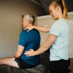 Warm, professional image of a physiotherapist working with a patient in a home setting, showing gentle neck assessment or manual therapy.